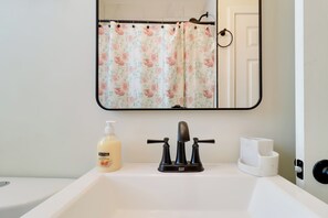 Een badkamer met een witte wasbak, een spiegel, een douchegordijn met bloemenprint en een zeepfles.