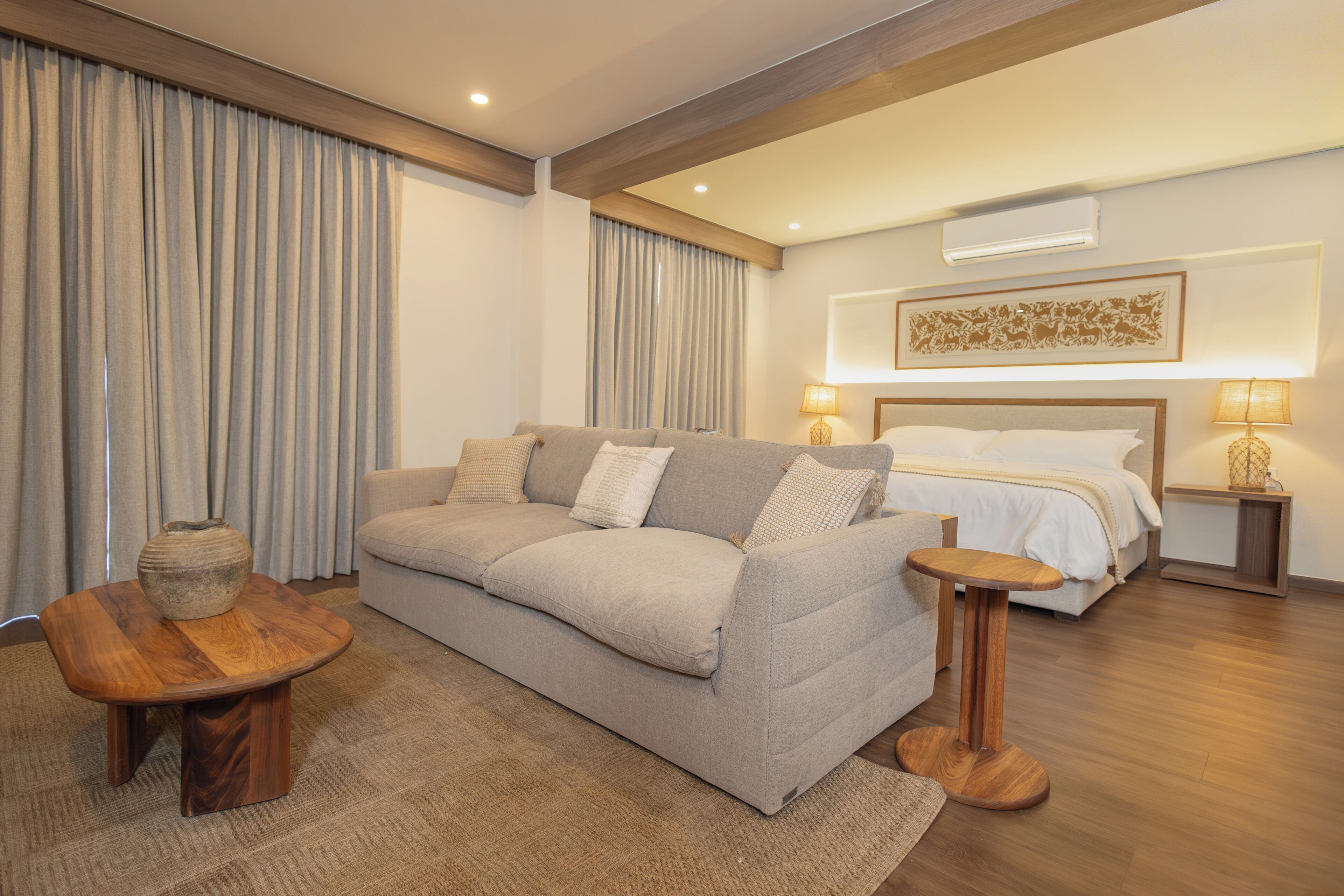 Una habitación de hotel con un sofá, una mesita de madera, una cama con edredón blanco y un aire acondicionado de pared.