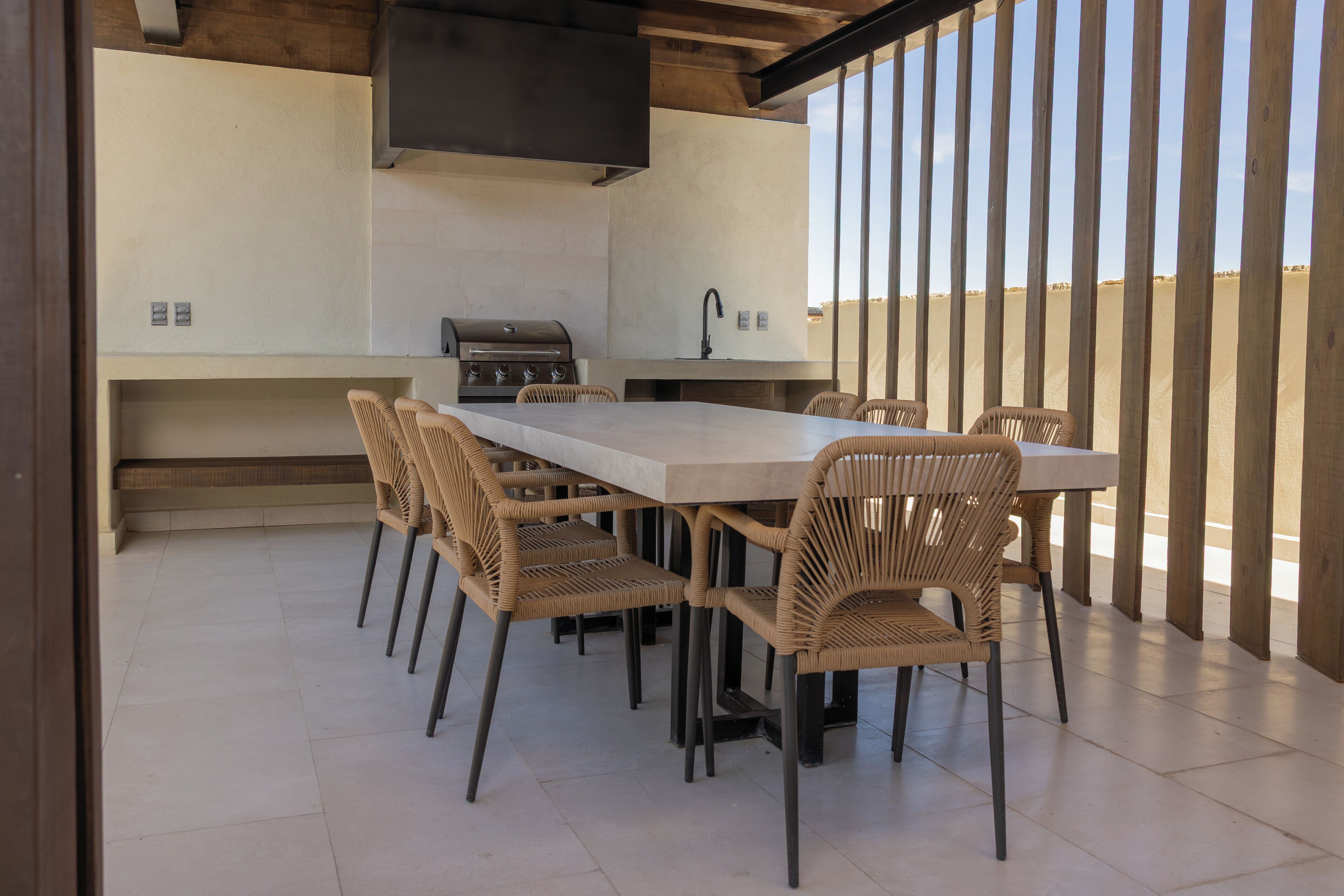 An outdoor kitchen with a long table and wicker chairs, a grill, and a view of the desert.