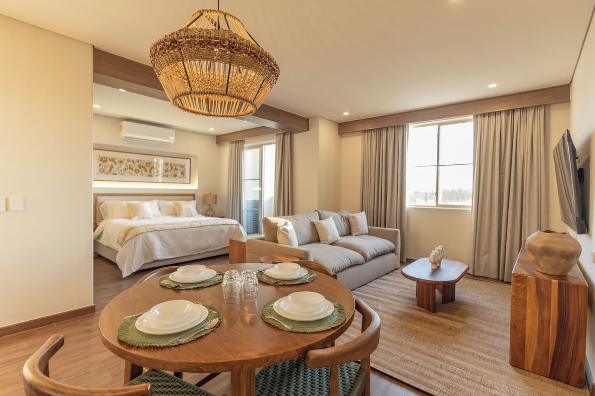 A hotel room with a dining table set, a sofa, a bed, and a large window.