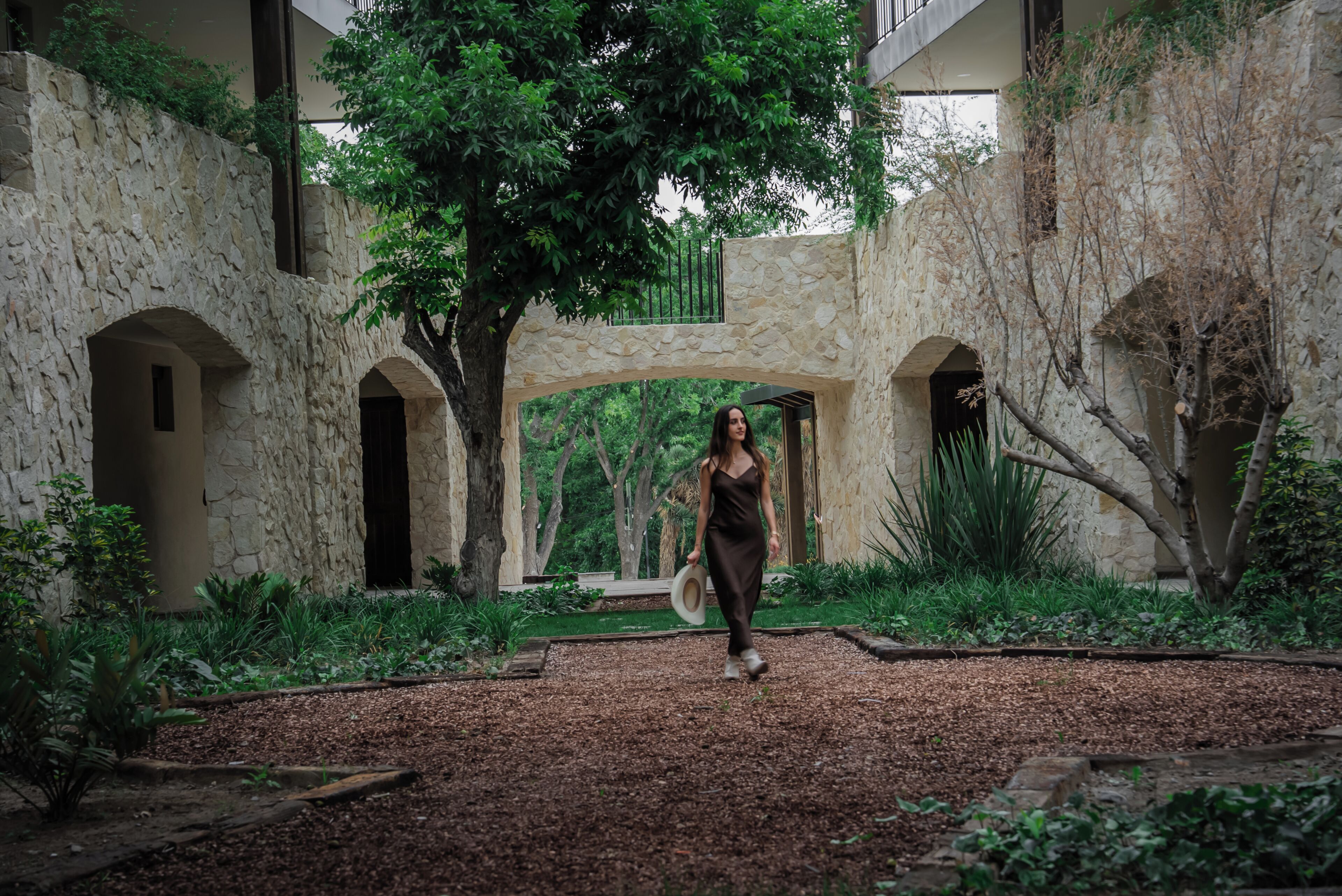 A woman walking through a courtyard with stone walls and a tree.