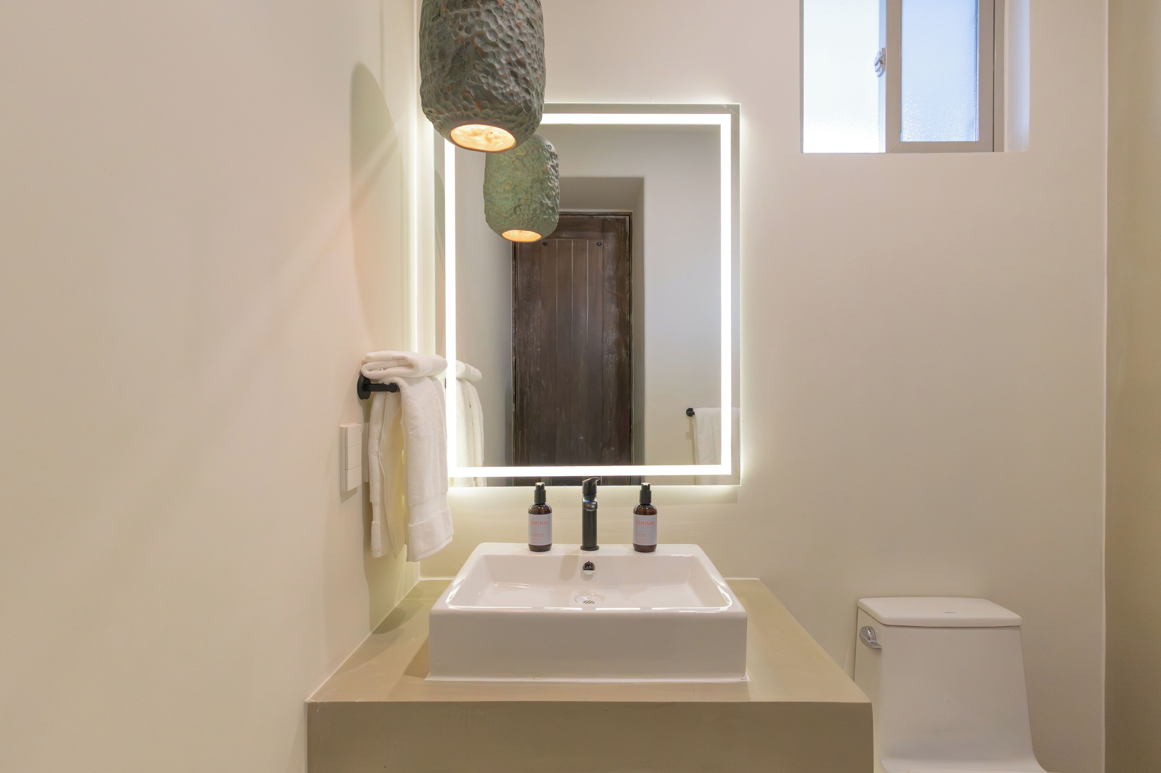 A bathroom with a rectangular sink, two bottles of soap, a towel, and a mirror with a light.