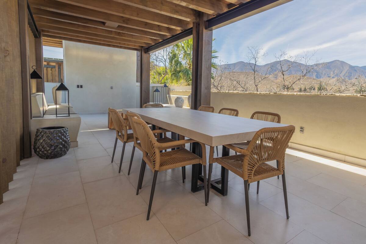 An outdoor dining area with a long table and wicker chairs, overlooking a mountainous landscape.