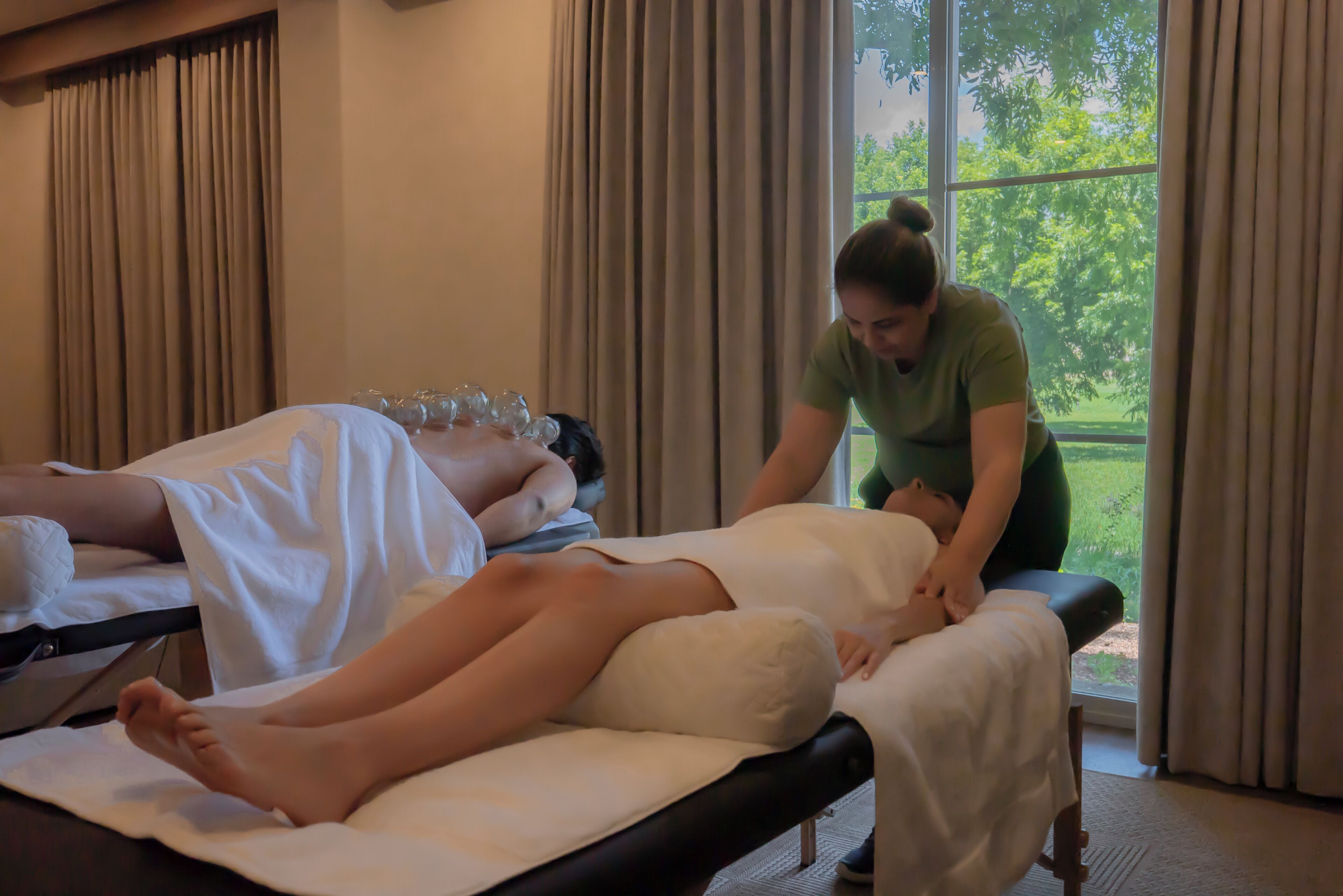 Un salón de masajes con dos clientes acostados en camillas de tratamiento, un terapeuta atendiendo a uno y un ventanal con vistas a la vegetación.