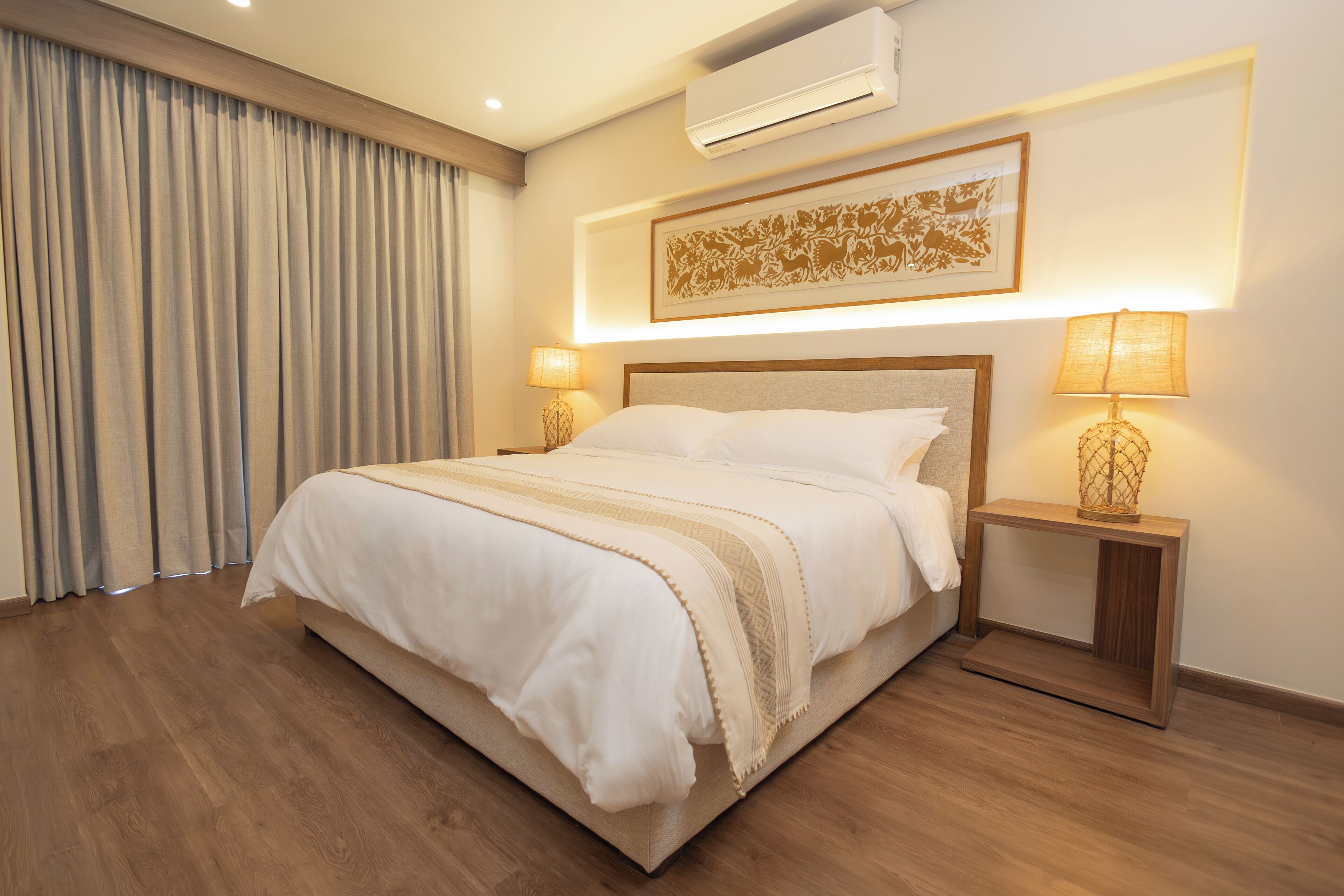 A hotel room with a large bed, two bedside tables with lamps, an air conditioner, and a framed artwork on the wall.