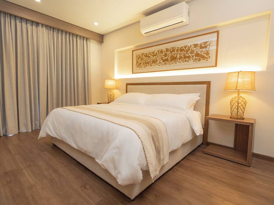 A hotel room with a large bed, two bedside tables with lamps, an air conditioner, and a framed artwork on the wall.