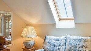 Ein Zimmer mit schräg verlaufender Decke, einer mit floralem Stoff bezogenen Couch, einer Lampe auf einem kleinen Tisch und einem Fenster mit Blick auf den Himmel.