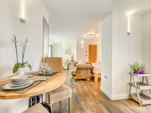 A modern dining area with a wooden table, chairs, and a vase of white orchids. There is a sofa and a small side table with a lamp in the background. - Luxurious, 5-star, open plan, ground floor apartment designed for two guests. (Bude)