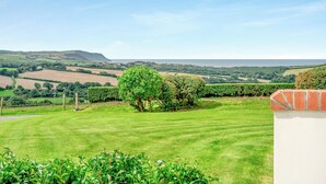 Ein grüner Rasen mit einer Hecke und einer Ziegelmauer im Vordergrund, mit Blick auf eine ländliche Landschaft mit Feldern und einer entfernten Meeransicht.