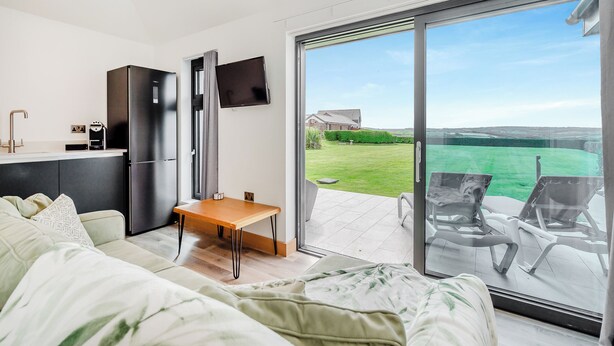 Ein modernes Wohnzimmer mit einer Couch, einem Couchtisch und einem an der Wand montierten Fernseher. Eine Glastür öffnet sich zu einem Balkon mit Sitzgelegenheit im Freien.