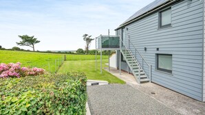A modern building with a grey exterior, a staircase leading to a covered outdoor area, and a well-maintained lawn with pink flowers. - Luxurious, 5-star, open plan, first floor apartment designed for two guests. (Bude)