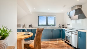 A modern kitchen with a dining area, featuring a large window, dark blue cabinets, and a stainless steel range hood. - Luxurious, 5-star, open plan, first floor apartment designed for two guests. (Bude)