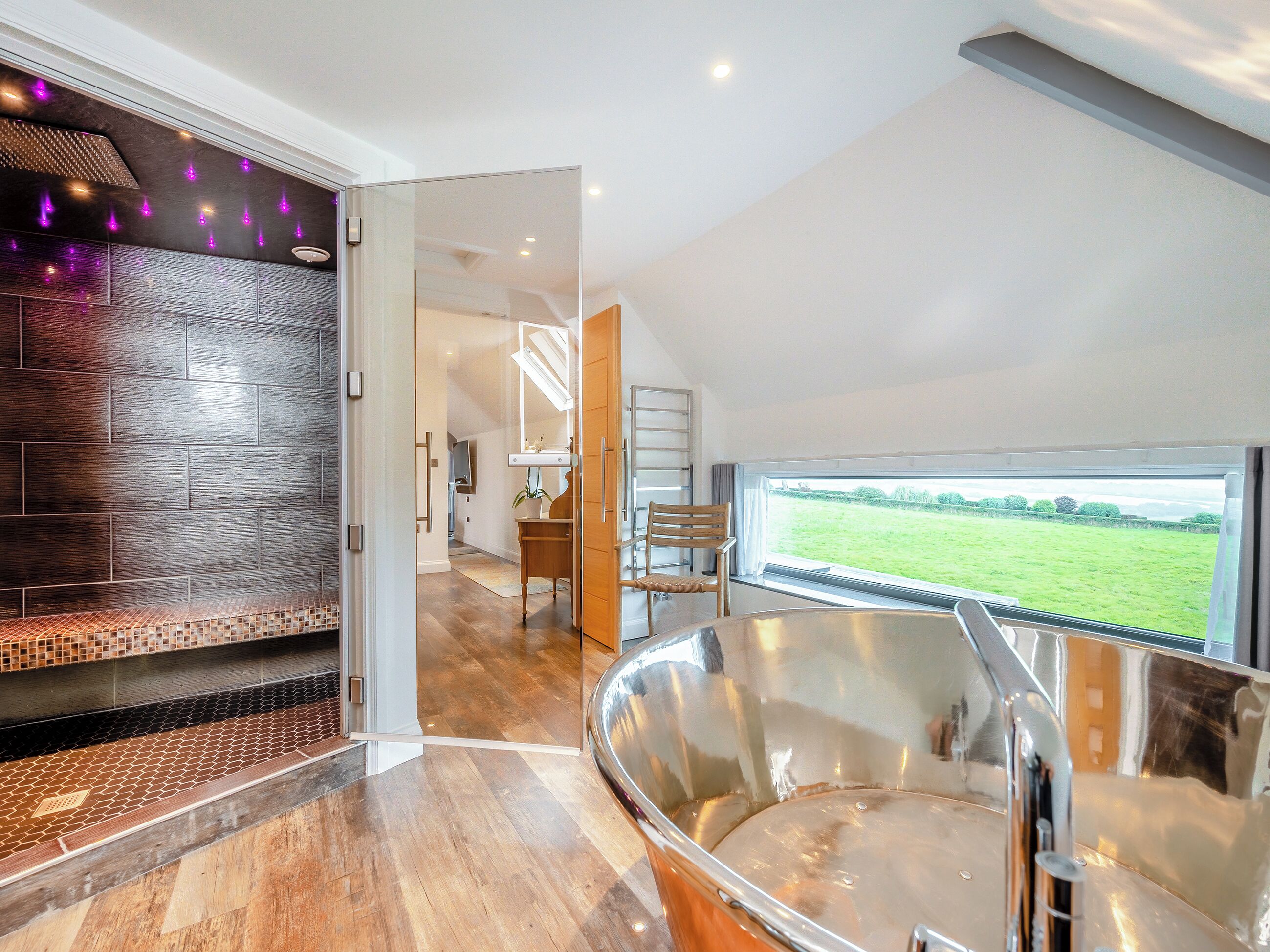 A modern bathroom with a large bathtub, a wooden bench, and a view of a green field.