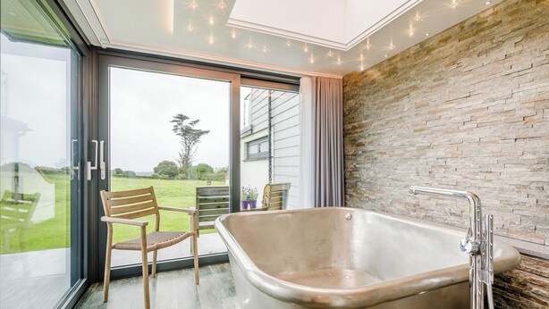 A modern bathroom with a large bathtub, a glass door leading to a balcony, and a view of the outdoors. - Sun Seeker - UKC6842 (Bude)