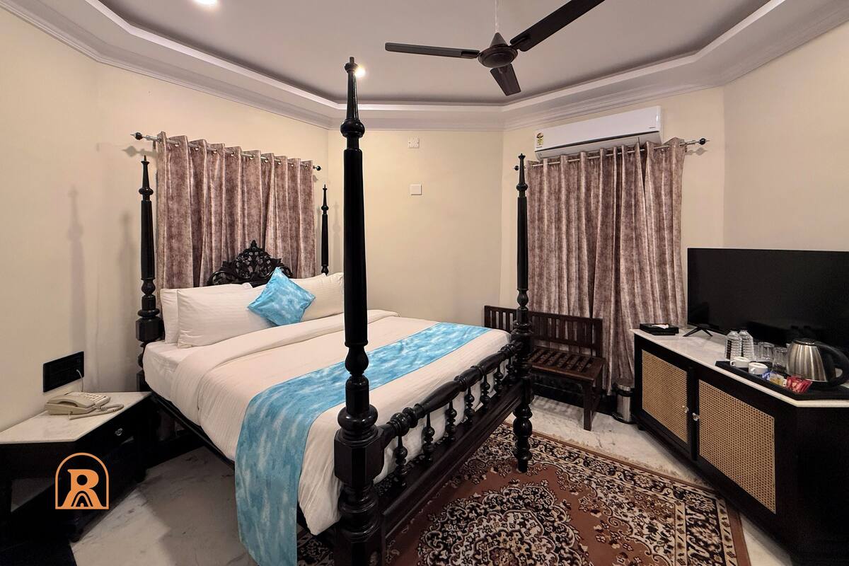 A bedroom with a four-poster bed, a ceiling fan, a television, and a small desk with a kettle.
