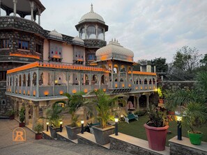 A traditional building with multiple balconies and a central dome, surrounded by potted plants and a paved courtyard. - The Shikargarh Palace (Udaipur)