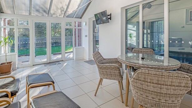 Patio soleggiato con tavolo in vetro e sedie in vimini, televisore montato a parete e vista sul giardino attraverso ampie porte scorrevoli.