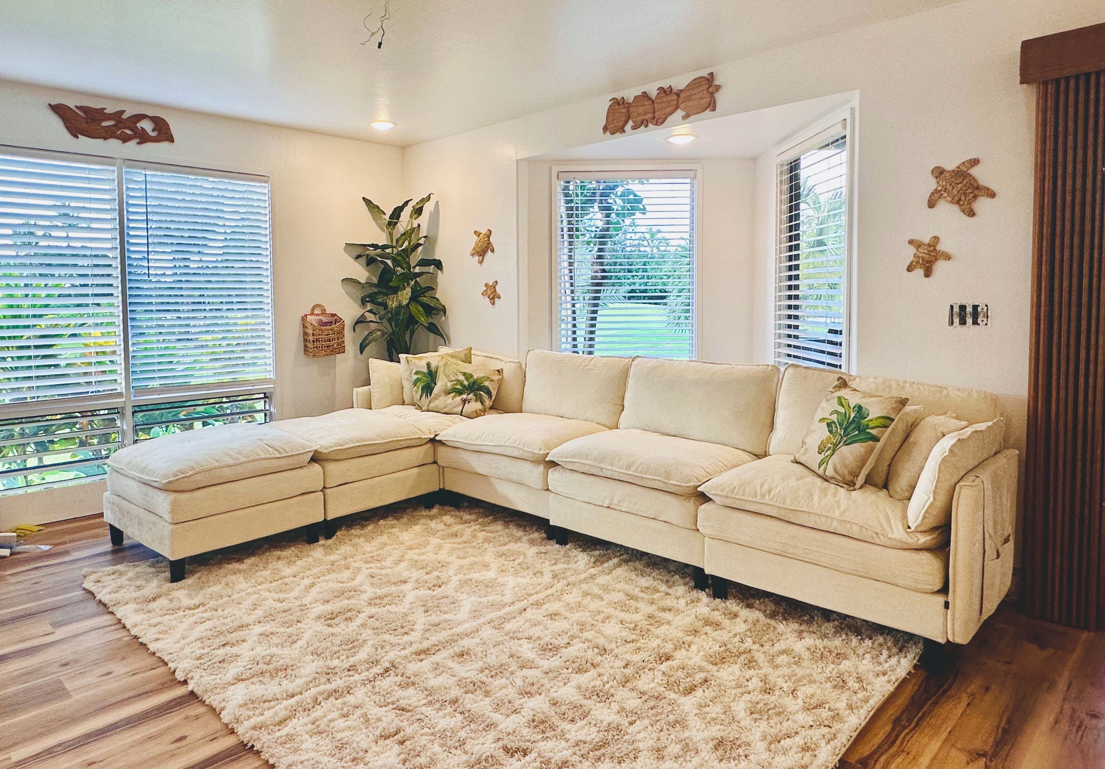 A L-shaped beige sectional sofa with a fluffy beige rug, two windows with blinds, a potted plant, and wall decor.