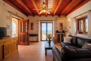 A spacious living area with a wooden ceiling, a ceiling fan, a television, a sofa, and a view through a glass door. - Villa Pi - Villa with a pool and splendid sea views (Vámos)