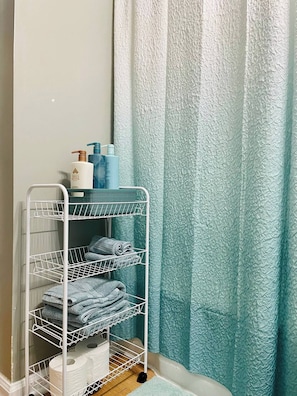A bathroom with a white metal rack holding towels and toiletries, a blue textured shower curtain, and a wooden floor. - Modern, Spacious, and Clean 2 Bedroom Townhome (Evansville)