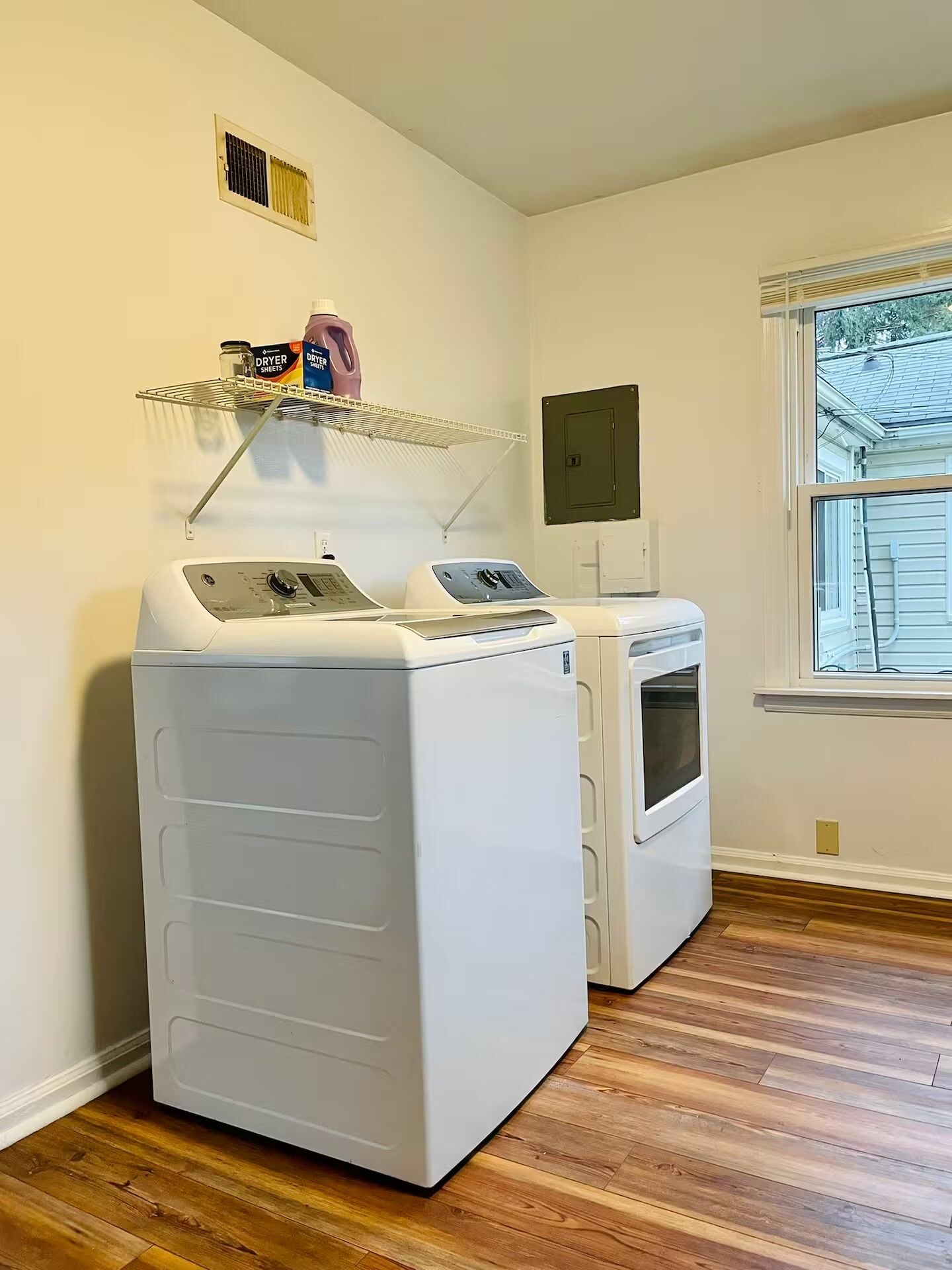 A laundry room with two stacked washers and dryers, a shelf with laundry supplies, a window with a view of the outside, and a door with a lock.
