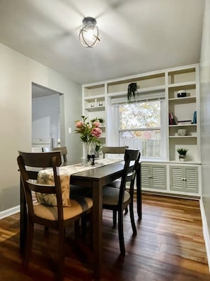 A dining area with a wooden table and chairs, a vase with flowers, and a window with shelves above. - Modern, Spacious, and Clean 2 Bedroom Townhome (Evansville)