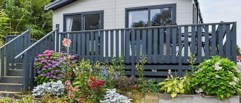 Ein Wohnmobil mit grauer Außenwand, einer Holzterrasse und einem Garten mit verschiedenen Pflanzen und Blumen.