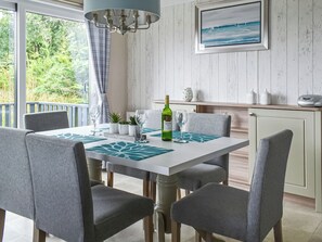 A dining area with a table set for four, chairs, a chandelier, a view of greenery outside, and a kitchen area with cabinets and a countertop. - Edmonton Lodge - UK50308 (Doublebois, near Liskeard)