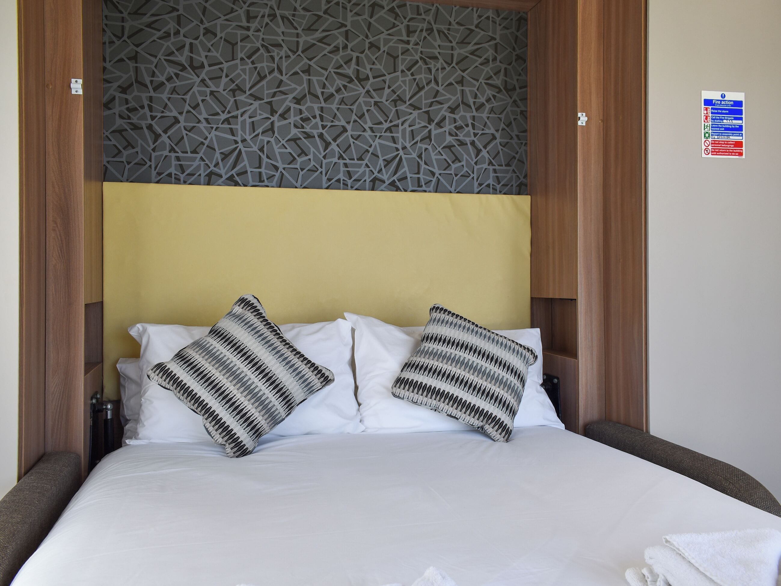 Een net opgemaakt bed met twee gestreepte kussens, een houten hoofdbord en een decoratief wandpaneel.