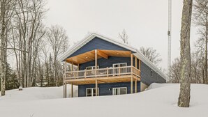 Una cabaña de dos pisos con balcón, rodeada de nieve en el suelo y entre árboles.