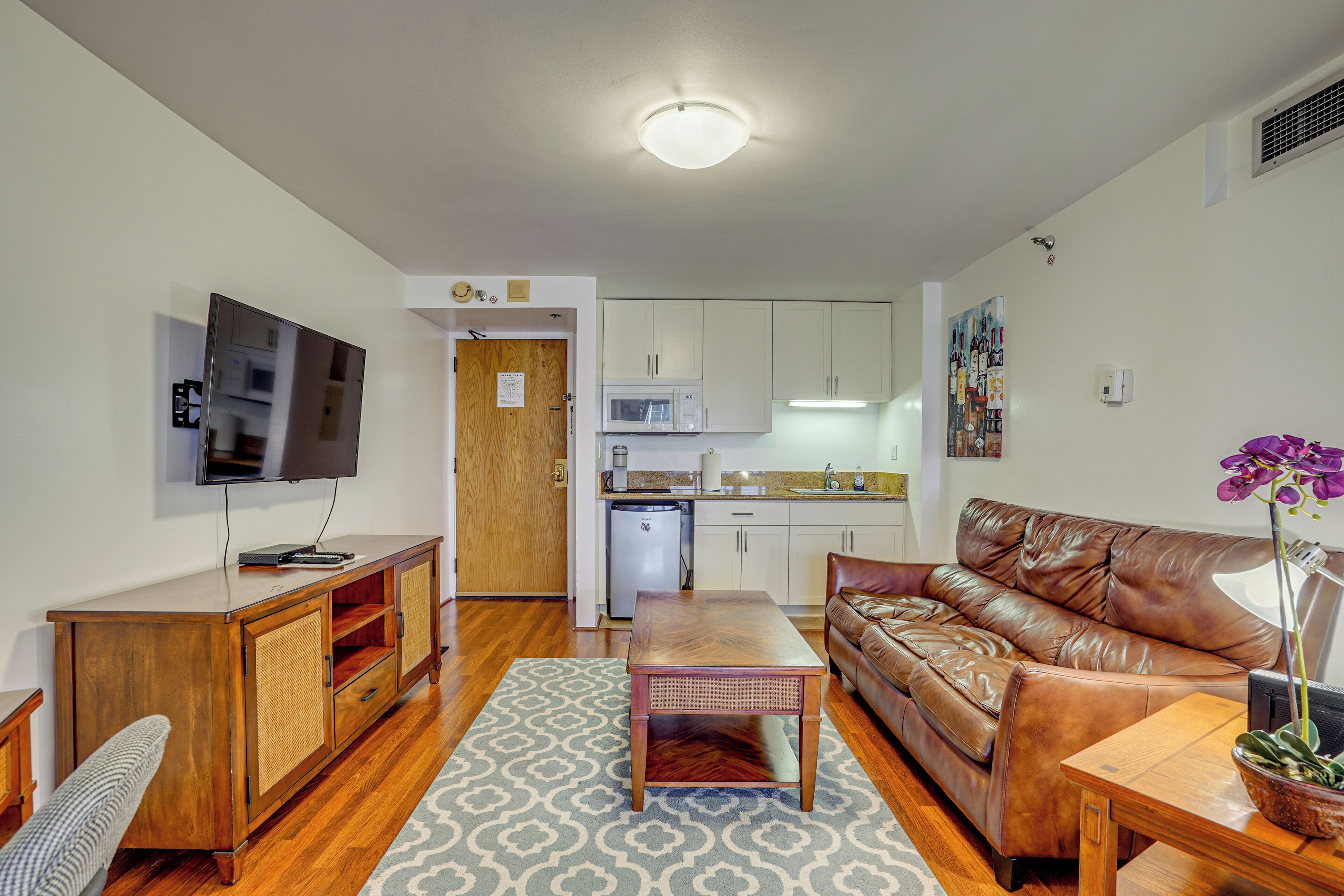 A compact living space with a wooden cabinet, a television, a brown leather sofa, a small table, and a kitchen area with white cabinets and a microwave.