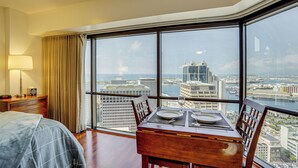 A hotel room with a large window offering a city view, a neatly made bed, a dining table set for two, and a lamp on a bedside table. - Free Parking Pool Harbor View Access Honolulu Gem! (Honolulu)