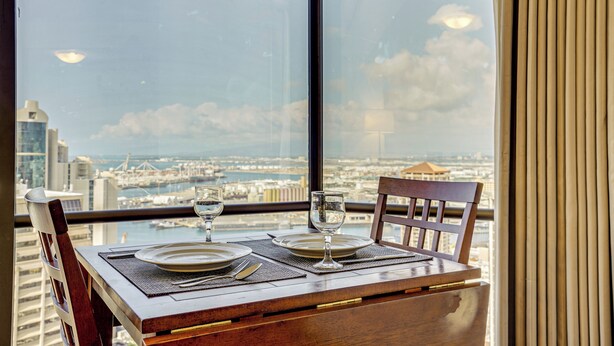 A dining table with plates, cutlery, and glasses set for two, overlooking a cityscape through a large window. - Free Parking Pool Harbor View Access Honolulu Gem! (Honolulu)