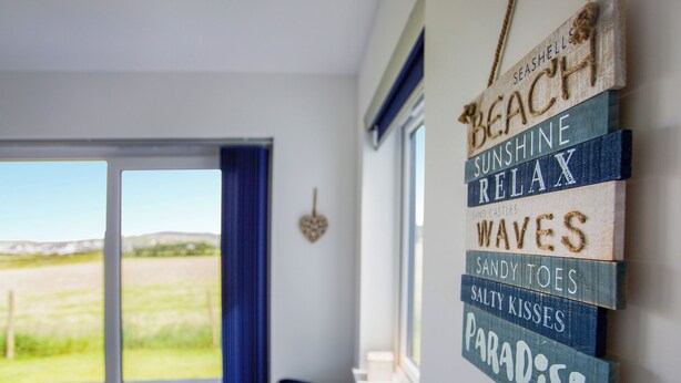 A room with a view of a green field through a glass door, a blue curtain, and a wall decorated with wooden signs reading "BEACH," "SUNSHINE," "RELAX," "WAVES," "SANDY TOES," "SALTY KISSES," and "PARADISE." - 4 bedroom accommodation in Blackwaterfoot (Blackwaterfoot)
