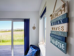 Habitación con vista a un campo verde a través de un vidrio, una cortina azul y una pared decorada con letreros de madera que dicen "BEACH", "SUNSHINE", "RELAX", "WAVES", "SANDY TOES", "SALTY KISSES" y "PARADISE".