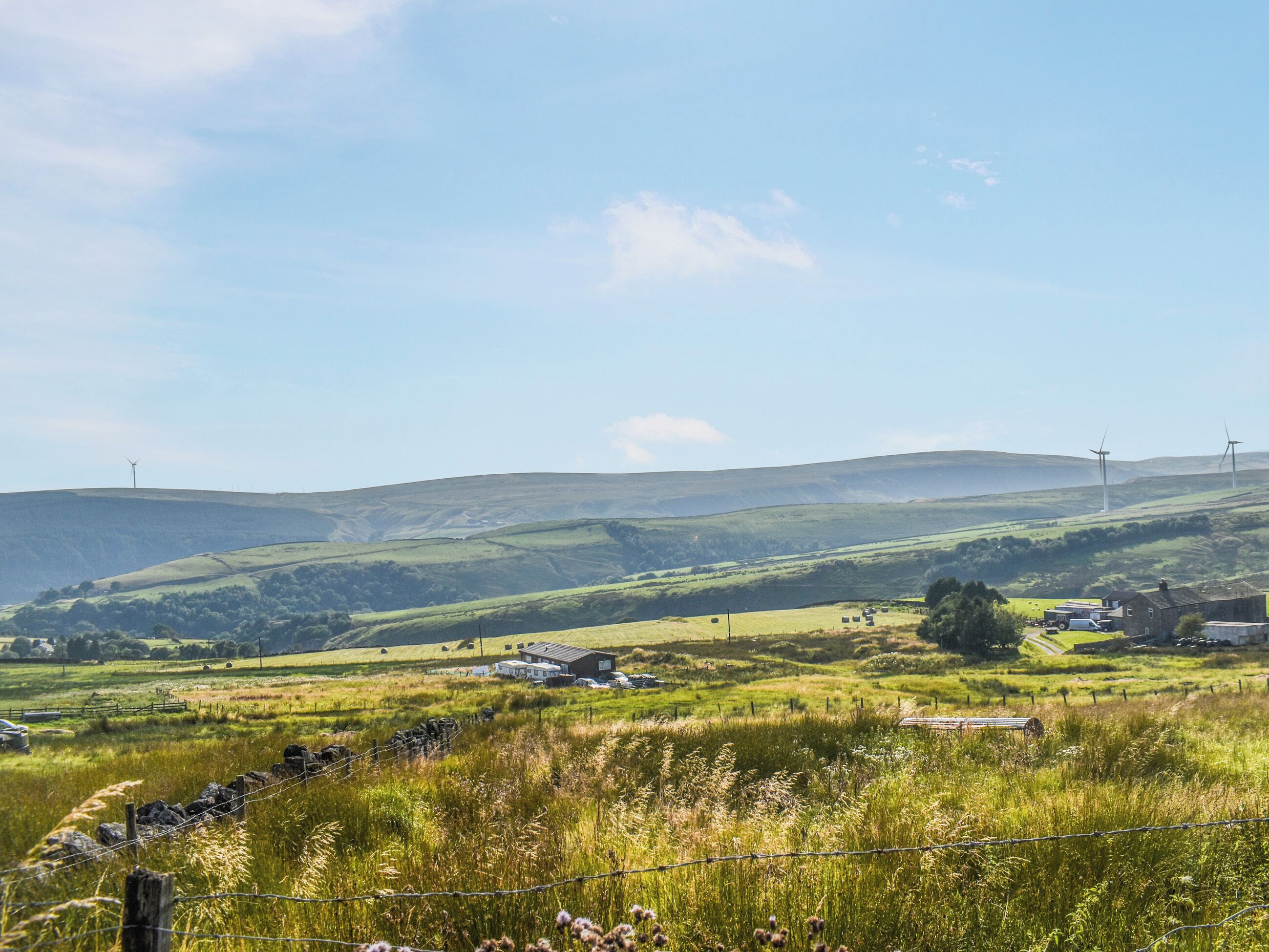 Ländliche Landschaft mit sanften Hügeln, einem Bauernhof und Windkraftanlagen in der Ferne.
