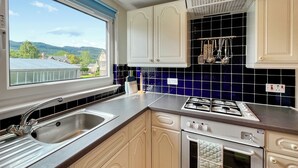 Une cuisine avec des meubles blancs, une cuisinière à gaz, un évier et une fenêtre donnant sur des maisons et des arbres.