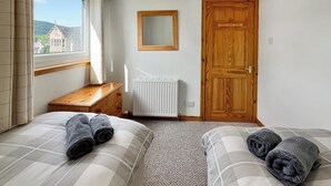 Une chambre avec deux lits, une commode en bois, un radiateur, une porte en bois et une fenêtre donnant sur des immeubles.