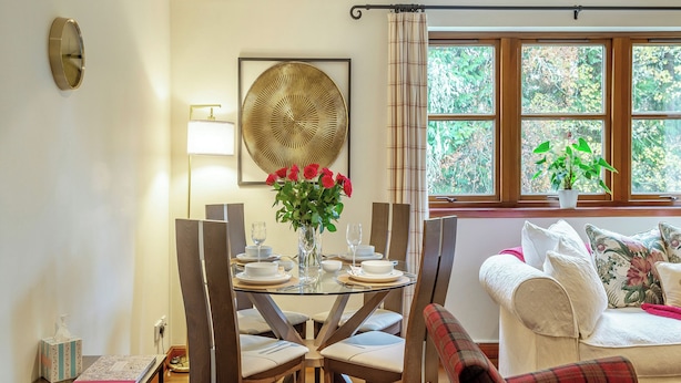Een eethoek met een ronde tafel voor vier personen, omringd door houten stoelen. Op tafel staat een vaas met rode rozen, en er is een raam met gordijnen achter.