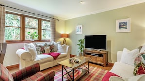 Een woonkamer met een bank, een geruite fauteuil, een salontafel, een televisie op een houten kast, en een raam met gordijnen.