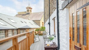Une terrasse en bois avec une table dressée pour le thé, un bâtiment en pierre avec un clocher en arrière-plan, et une pancarte indiquant « Castle 2 Cottage ».