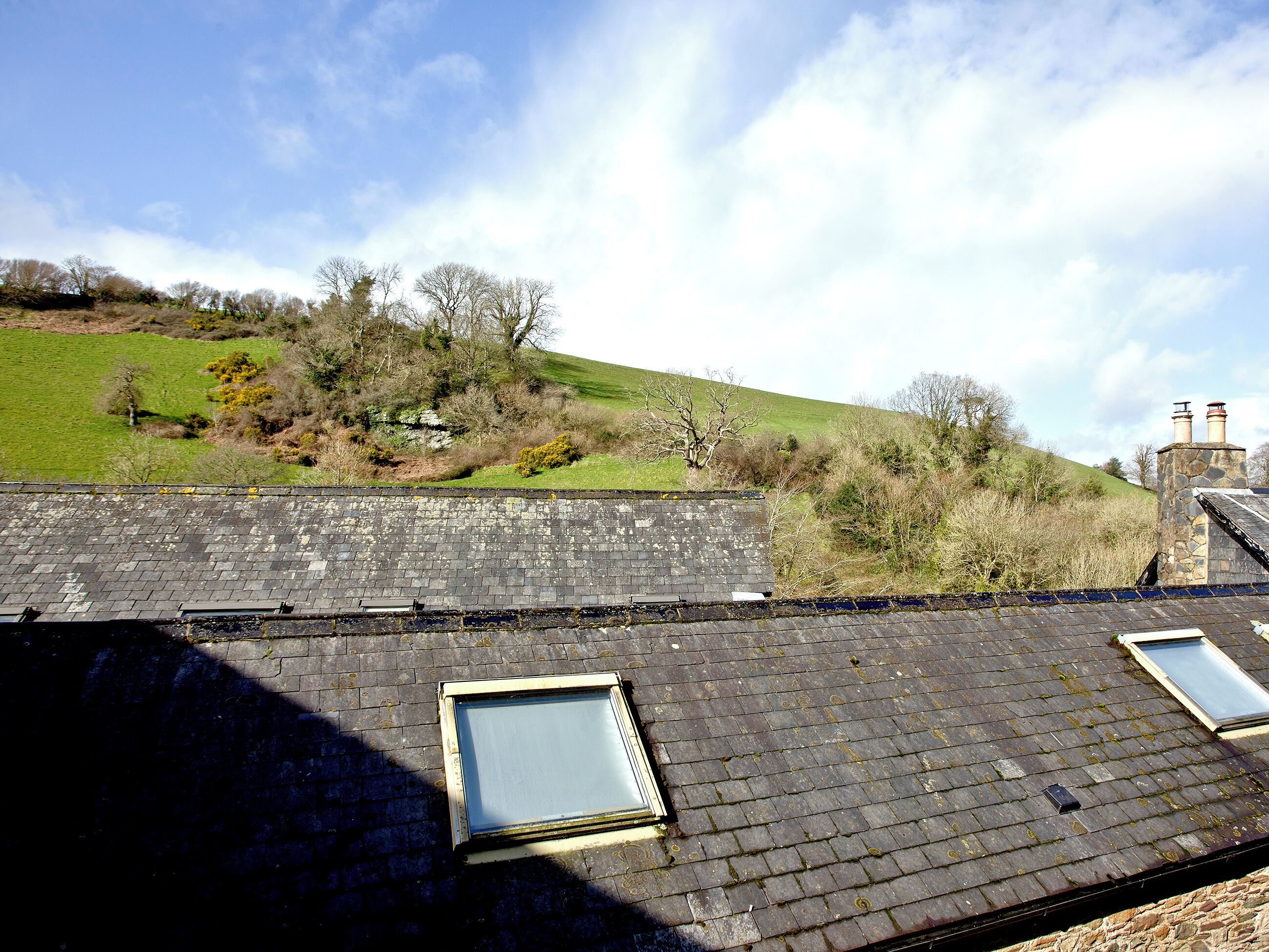 A rooftop with two skylights, a stone chimney, and a view of a green hillside.