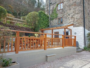 A wooden deck with a railing, a stone building in the background, and a white wall with a window. - Tuckenhay, 1 Castle cottage - FCK (Bow Creek, between Dartmouth and Totnes)