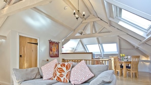 Un espacio tipo loft con techo alto, vigas a la vista, un sofá con cojines estampados, una mesa de comedor con sillas y una zona de cocina.
