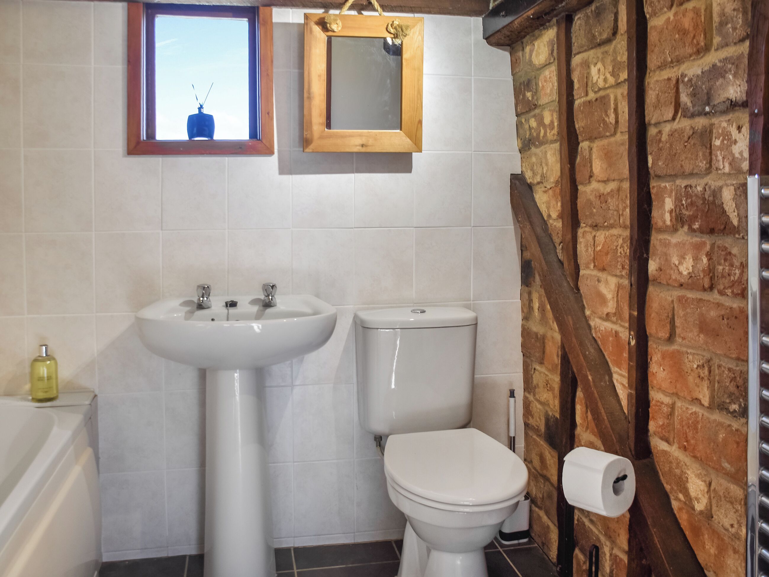 A bathroom with a white sink, toilet, and bathtub. There is a window with a view of the sky, a mirror, and a brick wall.