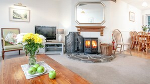 Una sala de estar con chimenea, televisor, zona de comedor con sillas de madera y una mesa con manzanas y flores.