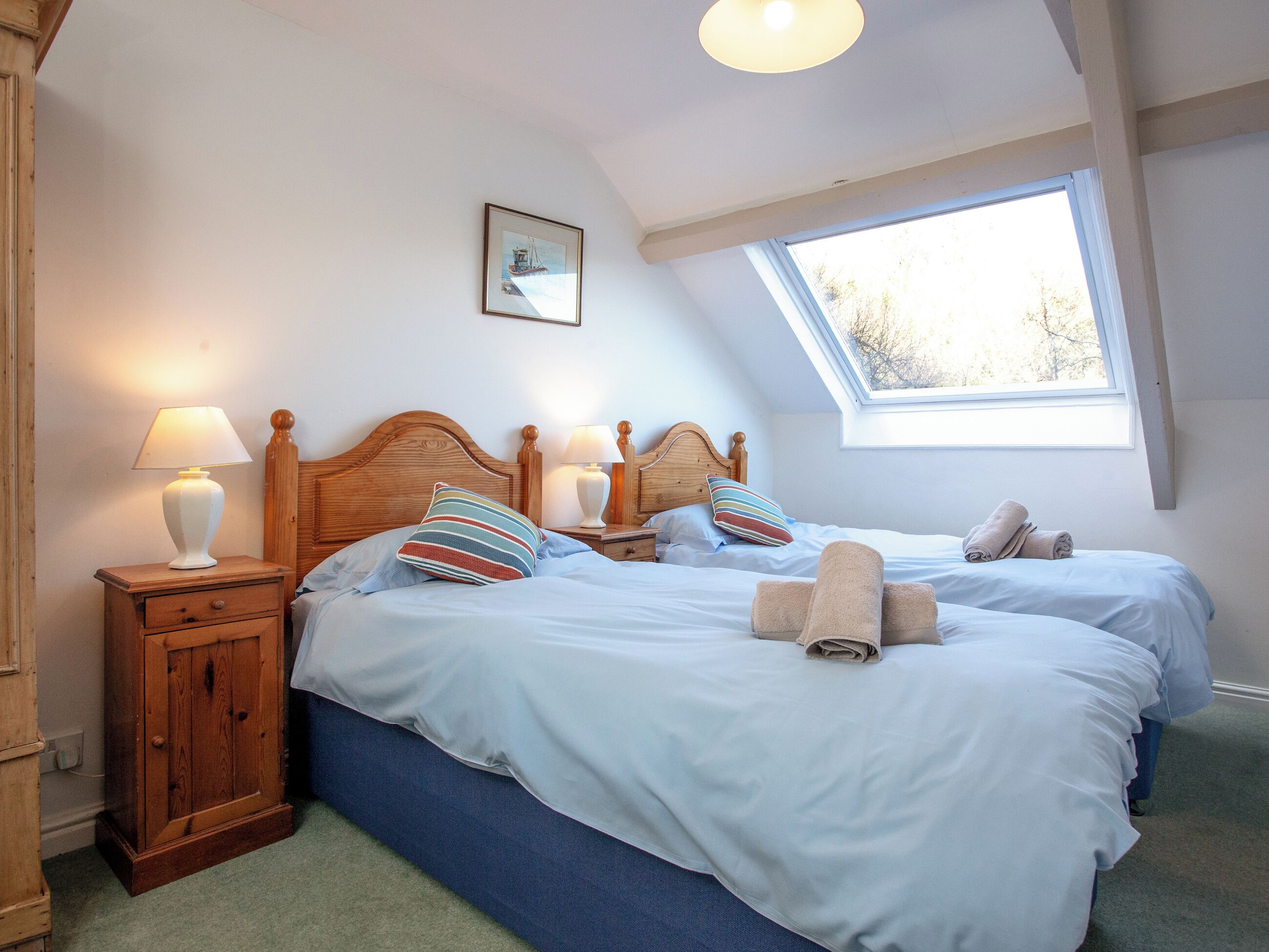 A bedroom with two beds, a wooden headboard, a nightstand with a lamp, a window, and a framed picture on the wall.