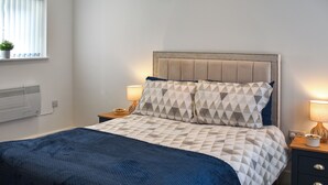 Un lit bien fait, avec une couette à motifs géométriques, une tête de lit capitonnée, des lampes de chevet et une petite table de chevet sur laquelle se trouve un bol.
