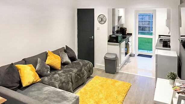 A modern living room with a grey sofa, yellow cushions, a yellow rug, a kitchen area with a sink, stove, and a small dining table. - Sunflower Bungalow - UK50569 (Little Dunham, near Kings Lynn)