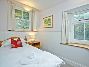 Een slaapkamer met een bed, nachtkastje, lamp, raam met gordijnen en een ingelijst schilderij aan de muur.